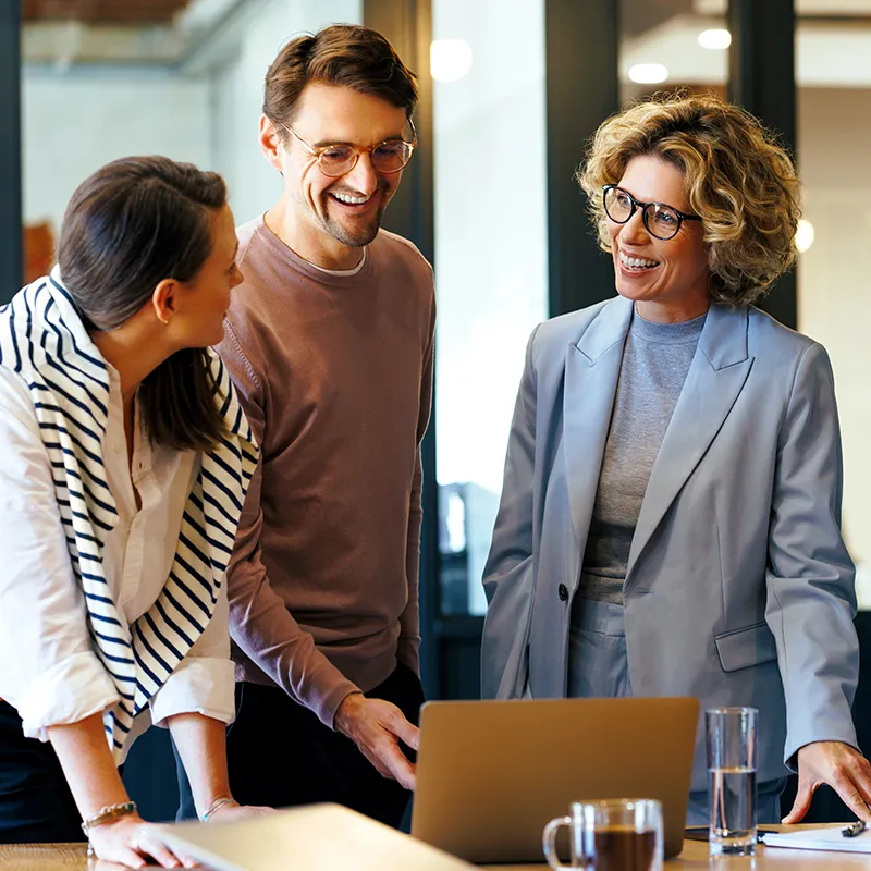 Two women and a man laughing together at work while looking at their computer.