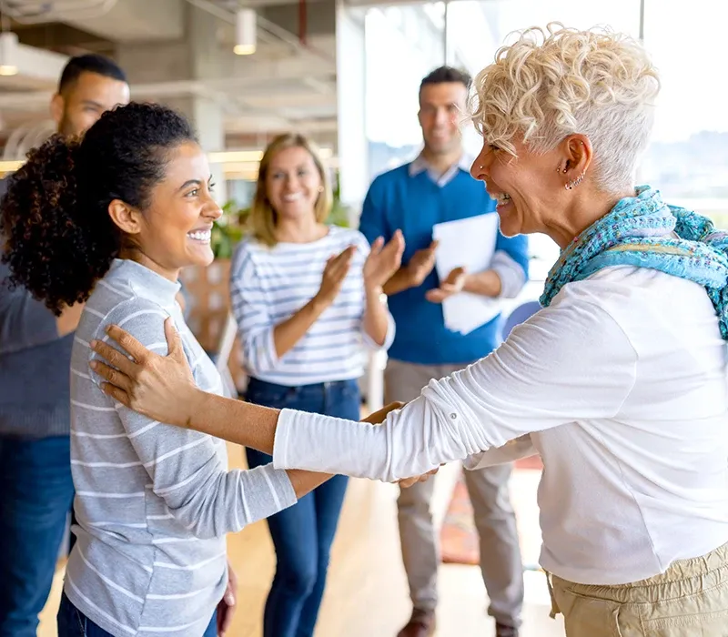 A woman congratulating a coworker while other team members applaud.
